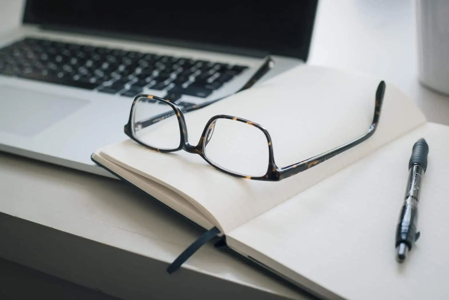 Eyeglasses on an open notebook next to a laptop and pen on a desk.